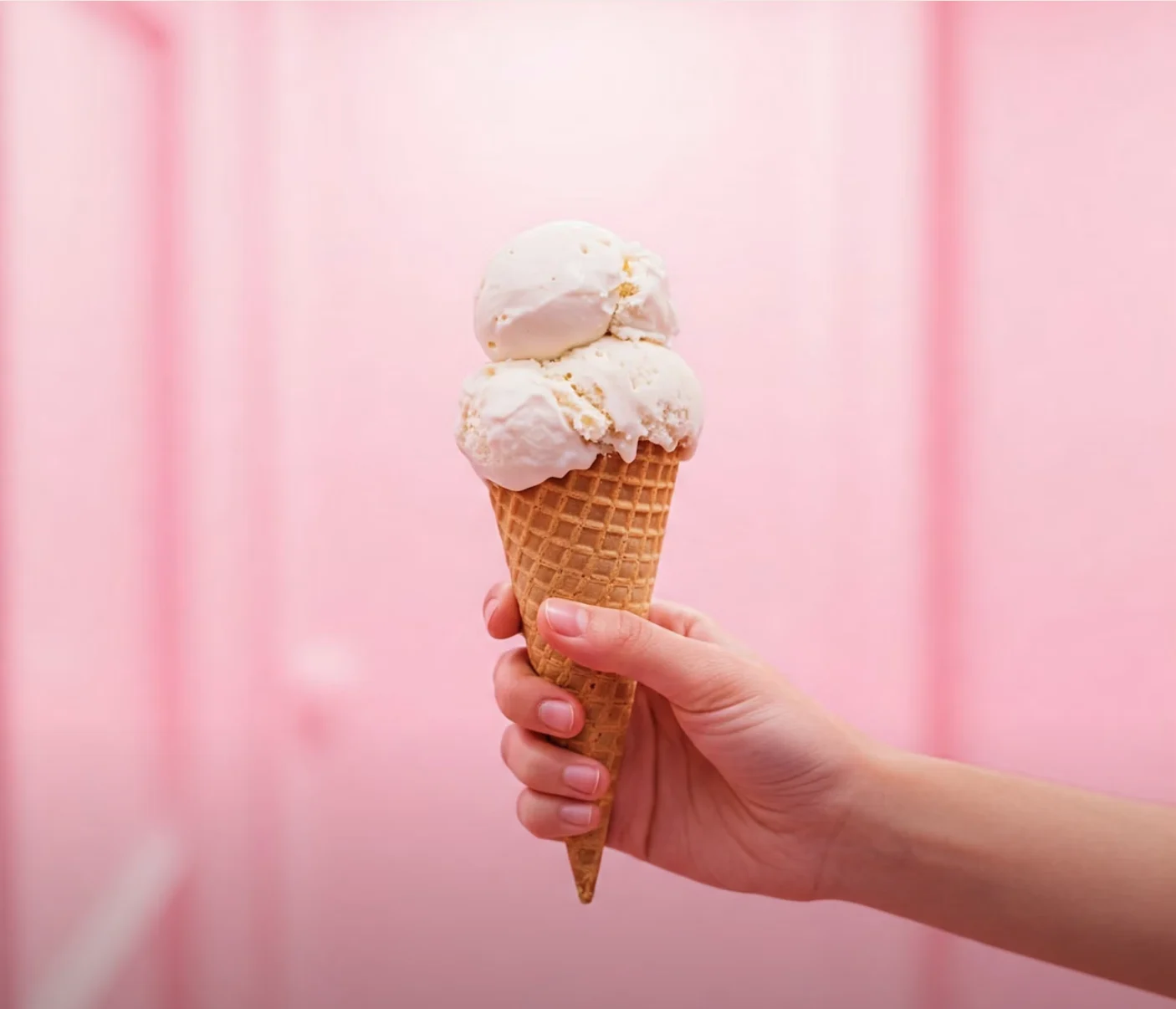 A hand holding a double scoop vanilla cone against a soft pink wall